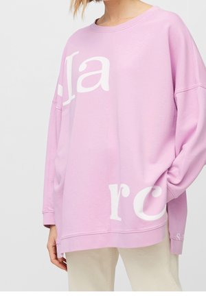 Kobieta w dużej jasnofioletowej bluzie z dużymi białymi częściowymi literami, w połączeniu z jasnobeżowymi spodniami, stojąca z ręką w kieszeni.