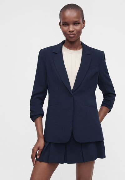 Femme portant un blazer bleu marine avec manches retroussées sur un haut blanc et une jupe plissée bleu marine assortie, sur un fond clair uni.