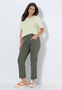 Mujer vistiendo una camiseta de manga corta color amarillo claro, pantalones verde oliva con bolsillos con cremallera y zapatillas blancas con detalles en naranja y amarillo.