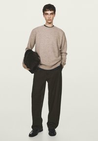 Maglione a maglia marrone chiaro, pantaloni larghi marrone scuro, scarpe nere, in mano una giacca scura. Design semplice, tessuto testurizzato, vestibilità rilassata.