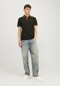 Donkergroene polo met oranje kraag en gestreepte mouwen, gecombineerd met lichtblauwe baggy jeans en witte sneakers.