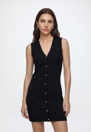 Femme aux longs cheveux bruns portant une robe noire sans manches avec des boutons dorés à l'avant et des boucles d'oreilles en anneau dorées, debout devant un fond uni.