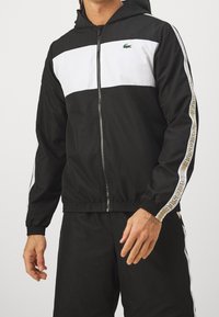 Man som bär en svart- och vit zip-up huvtröja med Lacoste-logotyp på bröstet och märkestripes längs ärmarna, samt matchande svarta byxor.
