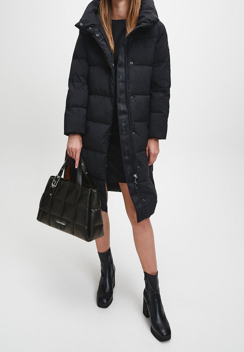 Femme portant un manteau matelassé noir longueur genou, des bottines noires, tenant un sac à main noir matelassé, debout devant un fond blanc uni.