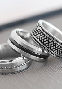 Vier metalen ringen op elkaar gestapeld: twee gladde zilveren banden, één met een zwart inlegwerk en één met een gestructureerd zwart diamanten patroon.
