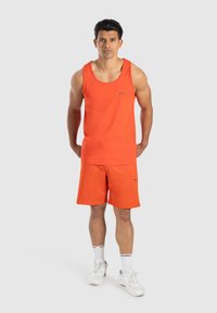 Oranssi tanktoppi ja shortsit, jotka on valmistettu pehmeästä kankaasta. Sisältää pienen logon rinnassa ja helmassa. Pukeutunut valkoisiin tennareihin ja sukkiin.