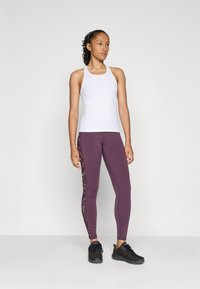 Vrouw in een witte mouwloze sporttop, paarse leggings met mesh zijkanten en zwarte hardloopschoenen, staand tegen een effen achtergrond.