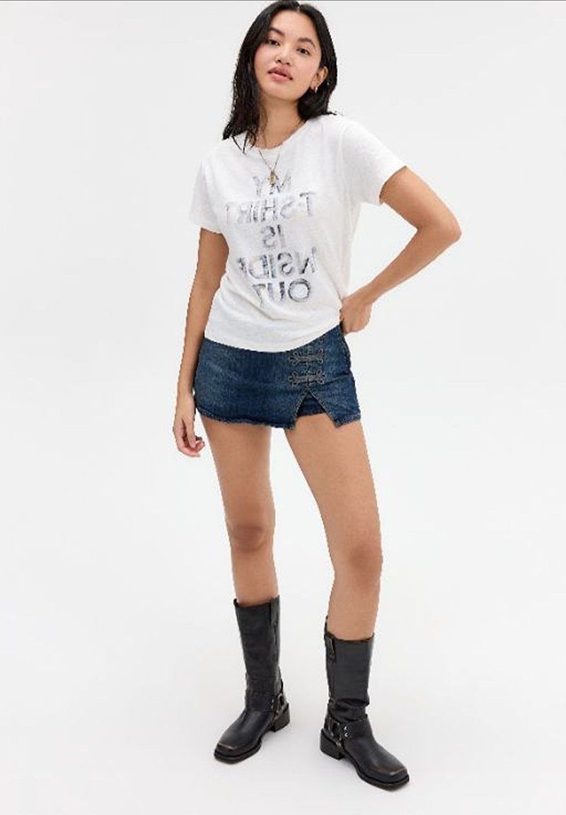 Νεαρή γυναίκα που φοράει λευκό t-shirt με σχέδιο, σκούρα μπλε μίνι τζιν φούστα και μαύρα μπότες μέχρι το γόνατο, στέκεται με το ένα χέρι στη μέση.