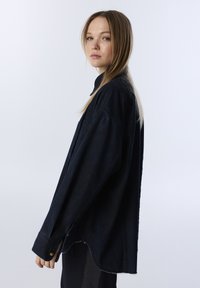 Camicia overshirt in denim scuro con una vestibilità ampia, maniche lunghe, polsini con bottone e un orlo leggermente curvo. Presenta un colletto e cuciture minimali.
