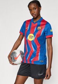 Φανέλα της FC Barcelona σε μπλε και κόκκινο με διαγώνιες ρίγες, κοντά μανίκια, V-λαιμόκοψη και κίτρινες λεπτομέρειες. Μαύρο σορτς Nike και μοτίβο σε ποδοσφαιρική μπάλα.