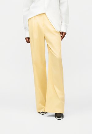 Woman wearing light yellow wide-leg trousers, white long-sleeve shirt, and black pointed shoes with silver tips, standing on white background.