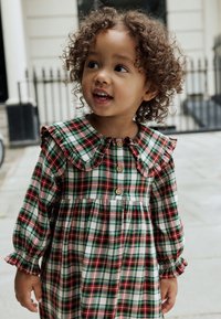 Robe à carreaux rouges, verts et blancs avec un col à volants, des manches longues et des boutons en bois sur le devant. Tissu doux en coton.