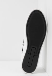 Semelle en caoutchouc noir d'une basket blanche montrant "MICHAEL KORS" en relief sur la zone du talon sur un fond blanc.