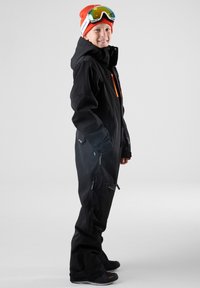 Schwarzer Schneeanzug mit orangefarbenen Akzenten, hohem Kragen, seitlichen und Beinreißverschlüssen, getragen mit einer orangefarbenen Mütze und Skibrille, zeigt ein Seitenprofil.