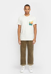 Crème kleur t-shirt met korte mouwen en een bergafbeelding op de linkerzak, gecombineerd met olijfgroene corduroy broek en witte sneakers.