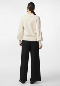 Frau mit langen Haaren, die mit einem schwarzen Band zusammengebunden sind, trägt einen cremefarbenen Pullover mit Puffärmeln und weite schwarze Hosen und steht vor einem weißen Hintergrund.