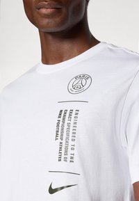 Biała koszulka z krótkim rękawem z czarnym i zielonym graficznym napisem. Posiada logo Paris Saint-Germain oraz emblemat Nike, wykonana z materiału bawełnianego.