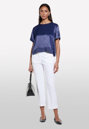 Top satin navy a maniche corte, design corto, fessure laterali. Abbinato a pantaloni bianchi a gamba dritta e ballerine nere, con una borsa nera in mano.