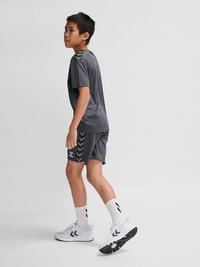 Jongen staat zijwaarts, draagt donkergrijze sportkleding, witte sokken en witte sneakers tegen een effen lichte achtergrond.