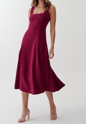 Kobieta w burgundowej sukience midi bez rękawów z kwadratowym dekoltem i rozkloszowanym dołem, nosząca beżowe sandały na wysokim obcasie, stojąca na białym tle.