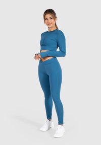 Türkisfarbener, kurz geschnittener Langarm-Top, kombiniert mit passenden Leggings, aus dehnbarem Stoff, mit figurbetontem Design und glatter Textur. Weiße Sneakers.