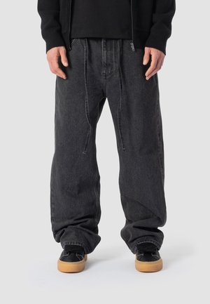 Person trägt locker sitzende dunkelgraue Jeans mit Kordelzug, schwarze Sneakers mit Gummisohlen und einen schwarzen Sweatshirt mit Reißverschluss.