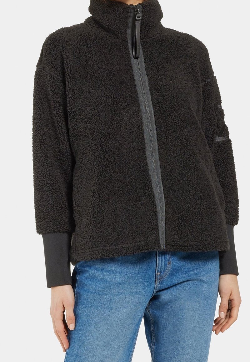 Personne portant une veste polaire noire avec une fermeture éclair verticale et un jean bleu, montrée du cou aux hanches sur un fond uni.