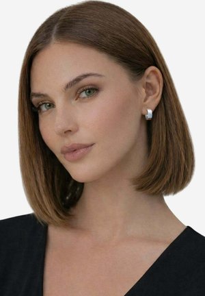 Donna con capelli castani lisci, che indossa orecchini a cerchio d'argento e una maglietta nera, che guarda leggermente di lato su uno sfondo uniforme.