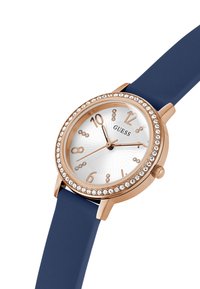 Rózsa arany színű óra kerek számlappal, fehér számlappal, kristály díszekkel, sötétkék szilikon szíjjal és aranyszínű órijelzőkkel.