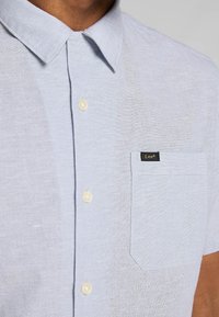 Camicia button-up in lino azzurro chiaro con colletto a punta, due bottoni e taschino sul petto con dettagli in etichetta nera.