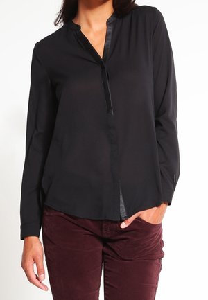 Blusa nera con colletto alla mandarino e patta frontale, caratterizzata da maniche lunghe e tessuto liscio. Indossata con pantaloni in corduroy bordeaux.