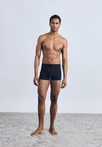 Μαύρα μαγιό σορτς με κορδόνι στη μέση, κατασκευασμένα από λείο υλικό. Το μοντέλο στέκεται ξυπόλητο σε μια ανάγλυφη, ανοιχτόχρωμη επιφάνεια.