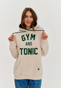 Béžová mikina s kapucí a zeleným nápisem "GYM AND TONIC", s tkaničkou, přední kapsou a žebrovanými manžetami. V kombinaci s modrými džíny.