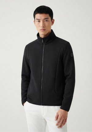 Młody mężczyzna w czarnej kurtce zapinanej na zamek z wysokim kołnierzem i białych spodniach, stojący na jednolitym jasnym tle.