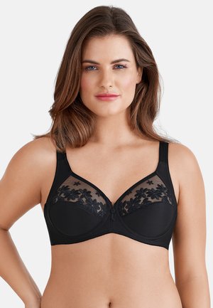 Frau mit schulterlangem braunem Haar, die einen schwarzen BH mit floraler Spitzenverzierung auf den oberen Körbchen vor einem einfarbigen Hintergrund trägt.