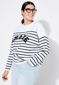 Felpa bianca a maniche lunghe con strisce orizzontali nere e la parola "AMAZE" ricamata in un carattere bold sul petto. Abbinata a jeans azzurri chiari.