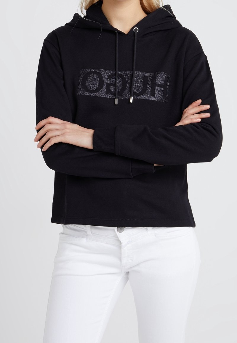 Femme portant un sweat à capuche noir avec un texte de marque scintillant et un pantalon blanc, bras croisés sur la poitrine, fond clair uni.