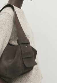 Ruskea mokkanahkainen olkalaukku, jossa on taitettava läppä ja näkyvä olkahihna. Pinta on teksturoitu ja siinä on minimalistiset metalliosat.