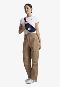 Borsa a tracolla blu navy con logo Converse, indossata a tracolla. Modello con una T-shirt bianca aderente e pantaloni cargo beige, con sneakers bianche.