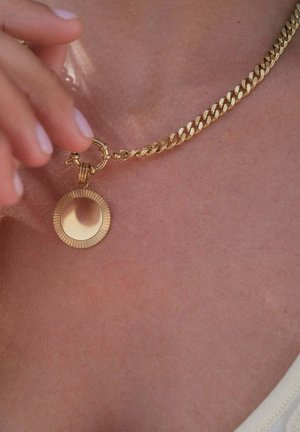 Hand mit weißem Ring hält eine goldene Kette mit rundem, strukturiertem Anhänger vor nackter Haut und weißem Stoff.