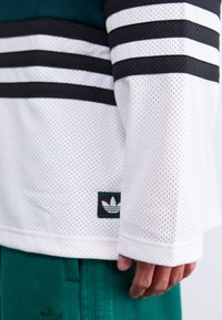 Osoba koja nosi bijelu mrežastu majicu dugih rukava s crnim prugama i Adidas logom, u paru s zelenim Adidas hlačama, ruka opuštena uz tijelo.