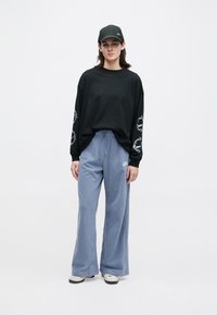 Mladý človek stojí v čiernej čapici a mikine s bielymi vzormi na rukávoch, modrých Nike teplákoch a bielych teniskách na svetlom pozadí.