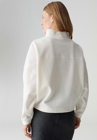 Weißes Sweatshirt aus Fleece mit hohem Kragen und überschnittenen Schultern. Glatte Textur und leicht gekürztes Design. Sichtbare einfache Nähte.