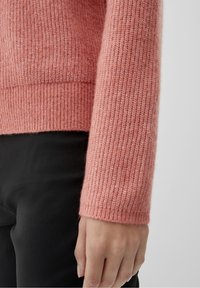Rosa stickad tröja med ribbade muddar och nederkant. Tyget verkar mjukt och något strukturerat, kompletterat av svart tyg nedanför.