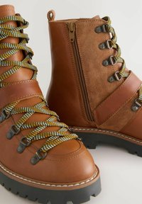 Braune Leder-Stiefeletten mit zweifarbigen Design, featuring grüne und gelbe Schnürsenkel, einem Reißverschluss und einer strukturierten Seitenpartie. Gummisohle mit Profil.