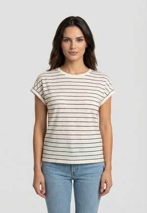 Donna con capelli lunghi e scuri indossa una maglietta bianca a maniche corte con strisce orizzontali nere e jeans azzurri, in piedi davanti a uno sfondo semplice.