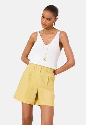 Žena nosící bílý tílkový top s výstřihem do V, žluté vysoké kraťasy s knoflíky, zlatý náhrdelník s příveskem a kruhové náušnice, stojící s rukama za zády.