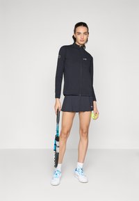 Marineblå hettejakke med teksturert stoff, kombinert med en matchende skort. Hvite sneakers med blå detaljer; holder en tenniskølle og ball.
