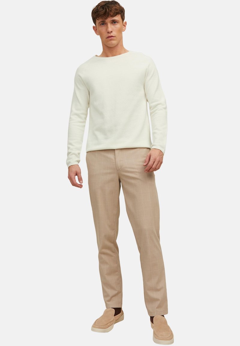Jeune homme debout portant un pull crème à manches longues, un pantalon à carreaux beige et des chaussures beige sans lacets, sur un fond uni.