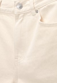 Zbliżenie na materiał dżinsowy w kolorze off-white, z metalowym guzikiem, zamkiem błyskawicznym i przednią kieszenią z widocznym ściegiem oraz gładką teksturą.